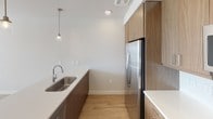 This single-floor space is designed with a clean, contemporary aesthetic that balances functionality and style throughout. The kitchen serves as a standout feature, showcasing flat-panel wood-toned cabinetry extending to the upper walls for generous storage, paired with crisp white countertops and a herringbone tile backsplash. Stainless steel appliances — including a French door refrigerator, electric range, over-the-range microwave, and undermount sink — complete the well-equipped cooking space. A pendant light fixture adds a refined touch above the sink area, while light wood-look flooring runs seamlessly throughout, contributing to a cohesive, warm interior.

The open layout connects the kitchen to a spacious living area, where multiple windows fitted with horizontal blinds allow natural light to pour in, casting bright patterns across the floor. The living space is finished with neutral gray-toned walls, recessed lighting, and a ceiling fan, creating a comfortable and airy atmosphere.

The bathroom offers a practical layout with a soaking tub and shower combination, white tile surround, and light wood-look flooring, maintaining the clean, minimal design language found throughout the rest of the space.