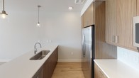 This single-floor space is designed with a clean, contemporary aesthetic that balances functionality and style throughout. The kitchen serves as a standout feature, showcasing flat-panel wood-toned cabinetry extending to the upper walls for generous storage, paired with crisp white countertops and a herringbone tile backsplash. Stainless steel appliances — including a French door refrigerator, electric range, over-the-range microwave, and undermount sink — complete the well-equipped cooking space. A pendant light fixture adds a refined touch above the sink area, while light wood-look flooring runs seamlessly throughout, contributing to a cohesive, warm interior.

The open layout connects the kitchen to a spacious living area, where multiple windows fitted with horizontal blinds allow natural light to pour in, casting bright patterns across the floor. The living space is finished with neutral gray-toned walls, recessed lighting, and a ceiling fan, creating a comfortable and airy atmosphere.

The bathroom offers a practical layout with a soaking tub and shower combination, white tile surround, and light wood-look flooring, maintaining the clean, minimal design language found throughout the rest of the space.