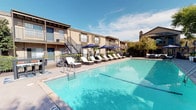 This single-story residence is situated within a well-appointed multi-unit community that offers notable shared amenities, including a large, sparkling swimming pool surrounded by a spacious sundeck furnished with lounge chairs and shaded by navy blue umbrellas. The building's exterior features a classic brick and neutral-toned facade with private balconies and stairways adding architectural interest.

Inside, the open-concept layout connects the living, dining, and kitchen areas in a cohesive flow. The living area is anchored by a fireplace with a mounted television above, flanked by large windows dressed in patterned curtains that admit generous natural light. Neutral gray walls complement the dark hardwood-style flooring throughout, creating a warm yet contemporary atmosphere.

The dining area accommodates a bar-height glass table and is brightened by multiple French doors leading to the outdoors, seamlessly blending interior and exterior living. A mirrored wall accent adds depth to this area.

The entryway features a decorative console table, mirrored wall art, and a large built-in storage closet. Windows throughout are fitted with horizontal blinds and layered curtains, offering privacy while maintaining brightness. The space carries a cohesive, modern aesthetic with teal and neutral accents throughout.