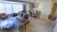 This single-floor residence offers a welcoming and functional layout designed for comfortable everyday living. The open-concept living and dining area serves as the heart of the space, where warm beige walls and soft wall-to-wall carpeting create a cozy, neutral backdrop throughout. The living area is generously sized, accommodating a plush sofa and accent seating, and is anchored by a large window dressed with vertical blinds that floods the room with abundant natural light. A sleek glass coffee table adds a modern touch to the seating arrangement. Seamlessly connected, the dining area features a round glass-top table surrounded by upholstered chairs, making it an ideal spot for meals and gatherings. Overhead, a contemporary track lighting fixture provides focused illumination, while a colorful abstract wall canvas adds a vibrant decorative accent. Decorative elements such as potted plants and woven storage pieces contribute warmth and texture to the overall aesthetic. The space maintains a clean, well-maintained appearance with consistent flooring and trim throughout, giving it a cohesive and polished feel. Overall, the layout balances open living with practical functionality in a bright and inviting atmosphere.