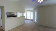 This single-floor space showcases an open-concept layout that seamlessly connects the kitchen and living areas. A defining architectural feature is the curved bay window area at the far end of the living space, which floods the room with natural light and lends a soft, rounded character to the floor plan. Multiple windows fitted with horizontal blinds allow for adjustable light control throughout the day.

The living area is finished with wall-to-wall neutral carpet, contributing to a warm and understated ambiance. A ceiling fan with a light fixture provides both ventilation and overhead lighting to the main space.

The kitchen is partially open to the living area via a raised pass-through counter, offering a functional connection between the two spaces without fully removing the sense of separation. White cabinetry lines the kitchen walls, complementing the light-toned countertop. Appliances are visible along the kitchen wall, including what appears to be a range and microwave unit.

The overall color palette of the space is neutral, with beige and off-white tones dominating the walls, flooring, and cabinetry, creating a clean and cohesive aesthetic throughout.