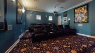 This dedicated home media room delivers an immersive, cinema-style experience from the comfort of a private setting. The room is finished with rich teal walls accented by crisp white crown molding, creating a dramatic yet polished atmosphere. A large projection screen dominates the front wall, flanked by wall-mounted surround sound speakers positioned for optimal audio coverage throughout the space. A ceiling-mounted projector anchors the viewing experience from above.

Seating is arranged in two rows of plush, dark leather recliner chairs — each equipped with cup holders — offering generous, theater-style comfort for multiple guests. The bold, patterned carpet featuring a colorful, playful design adds a lively contrast to the deep teal walls and dark furnishings.

The walls are adorned with framed Marvel Comics posters, including iconic covers featuring Iron Man and Captain America & Falcon, lending the room a fun, pop-culture character. A posted "Media Room Rules" sign and a wall-mounted AV equipment rack add functional, organized touches. Sconce lighting along the walls provides warm, ambient illumination, complementing the room's cinematic mood.

An adjacent entry area features light tile flooring and provides access to the media room through a standard interior door, offering a clean transitional space.