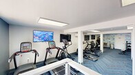 Located at 108 Summit Pointe Drive in Scranton, Pennsylvania, this fitness center offers a well-equipped, single-floor exercise space designed to support a full range of workout routines. The room is finished with a contemporary herringbone-patterned blue carpet that adds visual interest while providing a comfortable surface underfoot. Bright flush-mounted ceiling light panels illuminate the entire space evenly, creating a clean and energizing atmosphere.

The cardio zone features multiple treadmills, elliptical trainers, and a recumbent bike, each positioned for ease of use and accompanied by wall-mounted flat-screen televisions for entertainment during workouts. A dedicated free weights area includes a dumbbell rack with a full set of weights and an adjustable workout bench, catering to strength training needs. Additional resistance and cable machines round out the equipment offerings.

A bold, typography-style motivational mural covering an entire wall serves as a striking design focal point, reinforcing a wellness-oriented atmosphere. Fitness-themed artwork and posters further enhance the energetic ambiance throughout the space. Structural columns are integrated cleanly into the layout, and a modern cable-railing staircase provides access. A window near the entrance allows for a modest amount of natural light and an outdoor view, complementing the bright artificial lighting throughout.