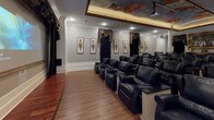 This private theater is a meticulously designed screening room that combines classical elegance with modern entertainment functionality. The main screening area accommodates a generous number of plush, black leather recliner seats arranged in tiered rows, each equipped with built-in cup holders and side tables, ensuring an optimal viewing experience from every position. Hardwood flooring runs along the base of the seating area, complementing the room's refined aesthetic.

The ceiling serves as a dramatic focal point, featuring a coffered tray design adorned with a hand-painted classical mural depicting celestial figures, framed by warm perimeter lighting. A large projection screen anchors the front of the room, framed by dramatic floor-to-ceiling black drapes with ornate gold-trimmed valances that echo throughout the space. Framed decorative wall panels with gilded botanical motifs are evenly spaced along the gray walls, reinforcing the theater's classical character. Crown molding adds architectural refinement throughout.

Adjacent to the screening room is a dedicated concession area, finished with rich wood cabinetry, a tiled floor with diamond accents, and a vintage-style popcorn cart. A gilded console table and ornate oil painting complete this lobby-like space, creating an immersive, grand cinematic experience.