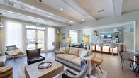 This thoughtfully designed single-floor community lounge offers an inviting and versatile environment suited for both casual relaxation and social gatherings. The open-concept layout seamlessly connects a comfortable seating area, a bar-style counter space, and a café-style dining nook, creating a cohesive and functional flow throughout the space.

The living area is anchored by a plush neutral-toned sofa and coordinating accent chairs, arranged around a substantial wood-and-stone coffee table. Decorative throw pillows and carefully chosen furnishings lend a warm, contemporary aesthetic. Adjacent to the seating area, a sleek bar counter with backlit shelving and high seating provides an ideal spot for socializing or working.

Architectural interest is added by the coffered ceiling with recessed lighting, which enhances the sense of depth and refinement. Large windows and glass-paneled French doors allow abundant natural light to pour in, offering views of an outdoor balcony area with greenery beyond. Sheer white drapery softens the light while maintaining an airy, open atmosphere.

The flooring transitions between warm wood tones and light tile, subtly defining each zone within the space. Overall, the lounge exudes a polished yet approachable ambiance, balancing modern design with everyday comfort.