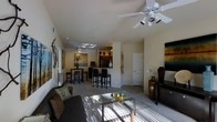 This single-story residence offers an open-concept layout that connects the living, dining, and kitchen areas into one cohesive and functional space. The living room is anchored by a plush dark sofa, a glass-top coffee table, and a flat-screen television, creating a comfortable gathering area. Ceiling fans in both the living and dining zones provide air circulation while contributing to the room's casual aesthetic.

A notable architectural feature is the vaulted ceiling with a skylight positioned above the dining area, allowing natural light to filter in and brighten the interior. The dining space is furnished with a dark wood table and matching bar-height chairs, complemented by a raised breakfast bar that transitions into the kitchen. The kitchen features dark cabinetry and appears to open toward the dining and living zones, reinforcing the open-plan design.

Neutral-toned walls and light carpeting run throughout, lending a clean and airy atmosphere to the space. Decorative wall art and carefully placed accents add visual interest without overwhelming the layout. The overall ambiance is relaxed yet polished, well-suited for both everyday living and casual entertaining.