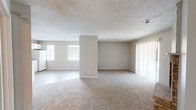 This single-story space offers a practical and comfortable layout with a neutral, move-in-ready aesthetic throughout. The open-concept living and dining area is expansive, featuring soft beige carpet, textured ceilings, and a central half-wall that subtly defines the two zones while maintaining an open, airy feel. Large vertical blinds along a sliding door allow natural light to flow into the space. A standout feature of the living area is the brick fireplace, framed by a chevron-patterned accent surround, adding a warm, classic focal point to the room.

The kitchen is efficiently designed with white raised-panel cabinetry extending to the ceiling, light granite-style laminate countertops, and light wood-look flooring. It includes an electric range with hood, dishwasher, and refrigerator, providing all essential appliances in a clean, cohesive white finish.

The bedroom is generously sized with plush carpet, neutral walls, and a ceiling fan with light fixture, offering both comfort and function. A door provides closet or hallway access.

The bathroom features tile flooring, a single-sink vanity with speckled countertop, and a tub-shower combination, finished in a clean, all-white palette that enhances the sense of brightness and cleanliness throughout.
