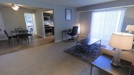 This single-floor space offers a practical and well-organized open-concept layout that seamlessly connects the main living area to the kitchen. The living area features wall-to-wall carpeting complemented by a textured area rug, providing a comfortable and inviting atmosphere. Large vertical blinds adorn the windows, allowing for adjustable natural light throughout the day. The space is set up to accommodate both relaxation and productivity, with room for a dedicated workspace complete with a desk and office chair alongside a central coffee table arrangement. The kitchen is accessible directly from the main living area and showcases stainless steel appliances, including a gas range with oven and an over-the-range microwave. A tile backsplash adds a modern touch to the kitchen, while dark cabinetry provides ample storage. The flooring in the kitchen transitions to hardwood-style flooring, offering a clean contrast to the carpeted living space. Neutral gray-toned walls throughout create a cohesive and contemporary aesthetic, making the space feel balanced and versatile. Overall, the layout is efficient and thoughtfully arranged, suitable for everyday living and working.