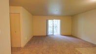 This single-floor residential space offers a clean, functional layout designed with comfort and simplicity in mind. The main living area is generously proportioned, featuring wall-to-wall carpeting in a neutral beige tone that adds warmth underfoot and complements the soft, light-colored walls throughout. The smooth ceiling overhead enhances the sense of openness, contributing to an airy and uncluttered ambiance. A standout feature of the living area is the sliding glass door that floods the space with natural light, framing a view of lush greenery beyond. The door opens to a private balcony enclosed by a classic railing, offering a pleasant outdoor retreat and a direct connection to the surrounding natural environment. The walls are painted in a warm, neutral hue that creates a calm and welcoming atmosphere, providing a versatile backdrop for a wide variety of interior styles. Multiple electrical outlets are conveniently positioned along the baseboards, ensuring practical usability throughout the room. Overall, this space presents a straightforward yet comfortable living environment, ideal for those seeking a bright, open, and low-maintenance home with a refreshing outdoor connection.