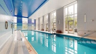Located at 300 Alexander Court in Philadelphia, PA, this expansive aquatic and wellness facility spans a single floor and offers a refined, resort-style environment for recreation and relaxation. The centerpiece of the space is a generously sized indoor lap pool, its crystal-clear turquoise water set beneath soaring double-height ceilings with a striking blue mosaic tile accent wall featuring horizontal stripe detailing in shades of blue, lavender, and white. Floor-to-ceiling windows line the pool area, flooding the space with abundant natural light while providing views of the surrounding cityscape and greenery. Gold ring fixtures punctuate the tiled wall, adding an elegant architectural touch. A separate, smaller plunge or whirlpool pool occupies an adjacent area, similarly adorned with vibrant blue mosaic tile work extending from floor to ceiling. A dedicated relaxation room offers two padded lounge chairs and a wooden side table, enveloped in soothing blue mosaic tile walls that create a tranquil spa-like atmosphere. The well-appointed locker room features rows of warm wood-toned lockers paired with a full-length mirror and a polished wooden bench, all set against neutral mosaic tile walls. Light tile flooring flows throughout, tying the various areas together into a cohesive, clean aesthetic.