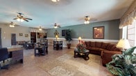 This single-floor space offers an open and versatile layout that seamlessly integrates a lounge area with a casual dining space. The living area is anchored by a plush brown sectional sofa paired with a dark wood coffee table adorned with floral arrangements, all set atop a patterned area rug over light tile flooring that extends throughout the space. Multiple ceiling fans provide comfort and airflow across the room.

The walls feature a two-tone treatment, with sage green on the upper portion and white wainscoting below, lending a clean, classic aesthetic. Framed artwork decorates the walls, adding warmth and character to the interior. A tall potted plant provides a natural focal point near the center of the room.

The dining area accommodates several small tables with dark wood chairs, suitable for casual gatherings or individual use. A striped canopy-style overhang marks the transition toward a background kitchen or service area. Textured popcorn ceilings and warm ambient lighting from table and ceiling-mounted fixtures contribute to the comfortable, relaxed atmosphere of the space.
