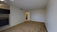 This single-floor space features a generously sized room finished with wall-to-wall beige carpeting, providing a neutral and versatile foundation suitable for a variety of furniture arrangements. The walls are painted in a clean, off-white tone that contributes to a bright and open atmosphere. A textured popcorn ceiling adds a classic architectural detail overhead, while a centrally placed HVAC vent ensures climate control throughout the space. A doorway along one wall opens into an adjoining room or hallway, where shelving units are partially visible, suggesting additional storage or utility space beyond. Electrical outlets are positioned at standard intervals along the baseboards, offering practical functionality throughout the room. The overall ambiance is clean, simple, and move-in ready, with a neutral palette that allows for easy personalization. The single-floor layout ensures convenient accessibility, and the straightforward design makes this space adaptable for use as a living area, bedroom, or flexible multi-purpose room.