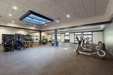 Located at 99 Gold Star Hwy in Groton, Connecticut, this single-floor fitness center offers a generously sized, open-plan layout designed to accommodate a full range of exercise activities. The space is organized into several distinct training zones, each thoughtfully equipped and finished to support an effective workout environment.

A standout architectural feature is the large, recessed skylight positioned prominently in the ceiling, flooding the central area with natural light and adding a sense of openness to the interior. Recessed lighting throughout the drop ceiling ensures consistent illumination across all zones. Durable rubber flooring in a dark, speckled finish covers the entire facility, providing safety and comfort underfoot.

The cardio zone lines one side of the facility and features treadmills, elliptical machines, and stationary bikes positioned beside a row of large windows with arched transom accents and decorative stained glass detailing, offering pleasant views of the surrounding greenery. A separate strength training area includes power racks, benches, free weights, and cable machines, with full-length mirrors along the wall to support proper form. An open functional training zone accommodates stability balls, a flat bench, and other accessories. A hydration station with a water cooler and small table is also present. The walls are finished in a combination of crisp white and deep navy blue accents, lending the space a clean, professional aesthetic.