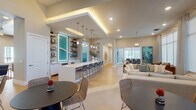 This thoughtfully designed single-floor space exudes a refined coastal aesthetic, blending modern architecture with warm, beach-inspired décor. The open-concept layout flows seamlessly between multiple functional zones, creating an inviting and social atmosphere throughout.

A central bar counter with a crisp white surface and decorative patterned tile base serves as the heart of the space, lined with woven strap barstools and illuminated by a row of elegant pendant lights. Behind the bar, built-in open shelving displays curated coastal accessories, flanking a striking backlit teal wave art panel that anchors the entire area with vibrant color and visual interest.

The lounge area features generous neutral sofas and wooden accent chairs arranged around coffee tables, bathed in abundant natural light streaming through expansive floor-to-ceiling windows dressed with sheer drapery. A bold abstract painting adds a splash of color to the relaxed seating zone, while views of palm trees hint at a warm outdoor environment.

Throughout the space, light wood-tone flooring, recessed lighting, and cove-lit tray ceilings contribute to the bright, airy ambiance. A distinct accent wall of horizontal shiplap boards, graduating from white to deep teal, reinforces the coastal theme. Additional dining nooks and a conference area provide versatile settings for both casual and formal gatherings, making this a fully equipped and visually cohesive communal destination.
