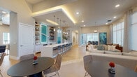 This thoughtfully designed single-floor space exudes a refined coastal aesthetic, blending modern architecture with warm, beach-inspired décor. The open-concept layout flows seamlessly between multiple functional zones, creating an inviting and social atmosphere throughout.

A central bar counter with a crisp white surface and decorative patterned tile base serves as the heart of the space, lined with woven strap barstools and illuminated by a row of elegant pendant lights. Behind the bar, built-in open shelving displays curated coastal accessories, flanking a striking backlit teal wave art panel that anchors the entire area with vibrant color and visual interest.

The lounge area features generous neutral sofas and wooden accent chairs arranged around coffee tables, bathed in abundant natural light streaming through expansive floor-to-ceiling windows dressed with sheer drapery. A bold abstract painting adds a splash of color to the relaxed seating zone, while views of palm trees hint at a warm outdoor environment.

Throughout the space, light wood-tone flooring, recessed lighting, and cove-lit tray ceilings contribute to the bright, airy ambiance. A distinct accent wall of horizontal shiplap boards, graduating from white to deep teal, reinforces the coastal theme. Additional dining nooks and a conference area provide versatile settings for both casual and formal gatherings, making this a fully equipped and visually cohesive communal destination.