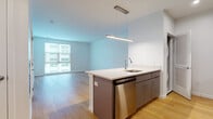 This contemporary single-floor unit is designed with a clean, modern aesthetic throughout. The open-concept layout connects the kitchen and living area seamlessly, creating a bright and airy atmosphere enhanced by large windows that invite abundant natural light. Light hardwood flooring extends throughout the main living spaces, contributing warmth and continuity to the design.

The kitchen is well-appointed with a central island featuring a built-in sink, stainless steel appliances including a refrigerator, range, microwave, and dishwasher, complemented by sleek gray cabinetry and white countertops. A modern pendant light fixture anchors the island as a focal point. A subway tile backsplash adds a refined finishing touch.

The open living area is generously proportioned, offering flexibility for various furniture arrangements. Large windows provide views of the surrounding exterior, further enhancing the sense of openness. The bedroom is a calm, well-lit space with the same hardwood flooring and a large window that contributes to its bright, uncluttered feel.

The bathroom is finished with light tile flooring, a combination tub and shower, a gray vanity with an undermount sink, and a large mirror, all contributing to a clean and functional design. Overall, the unit presents a contemporary and well-finished living environment.