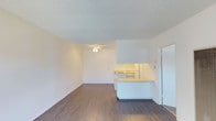This single-floor space offers a clean, open-concept layout that seamlessly connects the living area and kitchen. The living room is bright and airy, featuring warm wood-style laminate flooring that runs throughout the main area, complemented by crisp white walls that maximize the sense of light and openness. A ceiling fan is centrally positioned, providing both function and a subtle design element overhead. The textured popcorn ceiling adds a classic residential character to the space. The kitchen is defined by a raised breakfast bar counter that separates it from the living area while maintaining the open feel. White cabinetry lines the kitchen, offering ample storage, and under-cabinet lighting adds a warm glow to the workspace. The appliances are integrated neatly within the cabinetry. The layout is efficient and practical, making good use of the available floor space. Overall, the unit presents a neutral, move-in-ready palette with consistent finishes throughout, making it easy to personalize. The combination of wood-style flooring, white surfaces, and an open plan gives the space a simple yet inviting ambiance suitable for everyday living.