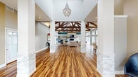 This fitness center and community clubhouse in Fort Worth, Texas offers a well-appointed single-floor facility spanning a generously sized footprint. The centerpiece of the space is a dramatic open lounge area defined by soaring vaulted ceilings with exposed dark wood truss beams and a striking floor-to-ceiling stone fireplace, above which a mounted television is anchored. Rich wood-look plank flooring runs throughout, complementing the stone-clad columns that frame the open layout. A wire-cage chandelier adds an elegant focal point upon entry, where a set of double glass doors welcomes natural light through an arched transom window. The lounge area is furnished with comfortable seating and workstation areas, creating a versatile communal environment. Large windows along the perimeter flood the space with daylight and offer views of the surrounding outdoors. The kitchen is fully equipped with dark wood cabinetry, white quartz countertops, a mosaic tile backsplash, stainless steel appliances including a dishwasher and microwave, and a center island — ideal for community gatherings. The dedicated fitness room features high ceilings, floor-to-ceiling mirrors, a full array of cardio machines, free weights, cable machines, and stability equipment, all set on durable flooring with ample natural light through surrounding windows.