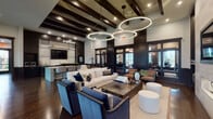 This elegantly designed single-floor amenity space offers a seamless blend of contemporary sophistication and warm, inviting interiors. Rich dark hardwood flooring runs throughout, complemented by dark wood trim, wainscoting, and dark-framed floor-to-ceiling glass doors and windows that flood the space with natural light while providing direct views of the landscaped outdoor courtyard and surrounding greenery.

The main lounge area is a standout, anchored by dramatic circular pendant light fixtures and exposed dark wood ceiling beams. Multiple seating arrangements — featuring plush sofas, velvet accent chairs, and sleek metal coffee tables — create an open yet intimate atmosphere ideal for gathering. A dedicated fireplace wall, clad in light stone tile and extending to a soaring ceiling height, adds a refined focal point, complete with a mounted television and tasteful decorative niches.

Adjacent to the lounge is a fully equipped kitchenette bar, featuring dark cabinetry, a light quartz-topped island with bar seating, pendant lighting, built-in appliances, and a mounted television — perfect for entertaining.

A dedicated business center provides two workstations with desktop computers, a printer, and a wall of dark built-in shelving, offering a productive and well-appointed workspace. Throughout the space, large sliding glass doors connect the interior to the outdoors, enhancing the sense of openness and connectivity.