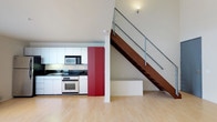 Located in the heart of San Diego at 320 West Ash Street, Unit 205, this two-story loft offers a modern, open-concept living experience. The main level showcases a well-appointed kitchen with white cabinetry, dark countertops, and stainless steel appliances, including a range and built-in microwave. A bold red accent panel adds a striking pop of color, creating a contemporary focal point. Light hardwood flooring runs throughout the main level, lending warmth and continuity to the space. Soaring ceilings amplify the sense of openness and volume, while abundant natural light enhances the airy atmosphere. A architecturally distinctive staircase with a rich wood stringer and sleek metal cable railing serves as both a functional and design element, leading to the upper level. The second floor overlooks the main living area below via a railed mezzanine, adding depth and dimension to the interior. The overall ambiance is clean and contemporary, blending industrial-modern influences with warm finishes to create a dynamic and versatile living environment well-suited to urban life in downtown San Diego.