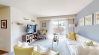 This well-appointed single-floor residence offers a thoughtfully arranged layout that balances comfort and functionality. The living room serves as the heart of the home, featuring soft carpeting, a soothing neutral color palette, and a striking blue accent wall adorned with framed artwork. A large sliding glass door floods the space with natural light and provides views of the surrounding exterior, while sheer white curtains add an airy, welcoming touch. The kitchen is efficiently designed in a U-shaped configuration, featuring rich dark wood cabinetry, light tile flooring, ample counter space, and a window above the sink that brings in additional natural light. The primary bedroom offers a calm and cozy retreat with plush carpet, a closet with paneled doors, and neutral walls that lend themselves to versatile décor. A secondary bedroom showcases built-in shelving and cabinetry surrounding the bed, offering generous integrated storage. The bathroom is finished with clean white subway tile surrounding a full soaking tub and shower combination. Throughout the residence, carpeted floors, consistent neutral tones, and natural light create a cohesive and comfortable atmosphere suited for everyday living.