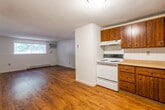 This single-floor unit offers a practical and functional layout across several distinct areas. The open living space is anchored by warm hardwood-style flooring that extends throughout the main areas, complemented by a textured ceiling and a large horizontal window that allows natural light to fill the room. A wall-mounted air conditioning unit and baseboard heating provide year-round climate control.

The kitchen is efficiently arranged in an L-shaped configuration, featuring rich dark wood cabinetry — both upper and lower — offering ample storage throughout. The kitchen is equipped with a white electric range, a white refrigerator, a black dishwasher, and a double-basin sink with tile backsplash lining the walls. Light-toned countertops provide functional workspace.

The bedrooms are carpeted and offer neutral wall tones, creating quiet and comfortable retreats. Each room features standard closet access via wooden doors. A textured ceiling is consistent throughout the private spaces.

The bathroom includes a full tub-shower combination surrounded by classic white square tile, giving the space a clean and timeless appearance. The entry area features hardwood-style flooring and a solid wood front door, providing a welcoming transition into the main living space.