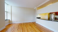 Located at 227 Church, this single-floor unit presents a clean, modern aesthetic throughout its well-designed layout. The main living area is anchored by warm-toned hardwood flooring that extends seamlessly across the space, complementing the neutral cream-colored walls and crisp white baseboards. Natural light filters in through a window fitted with blinds, brightening the open feel of the room. A structural column subtly defines the transition between the living area and the kitchen pass-through, which features a curved dark granite countertop overhang illuminated by under-cabinet lighting with a warm yellow glow.

The galley-style kitchen is efficiently organized, showcasing rich wood cabinetry paired with dark granite countertops and dark tile flooring. Stainless steel appliances, including a refrigerator, range, microwave, and dishwasher, lend a polished, contemporary character to the space. Under-cabinet lighting adds both functionality and ambiance.

The bathroom maintains a consistent design language with dark granite tile flooring and matching baseboard trim. A pedestal sink, full-width vanity mirror, and a row of modern light fixtures above contribute to a clean and functional finish. A tiled shower area completes the space.