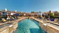 The outdoor amenity area at 6201 Wind Haven presents a well-appointed resort-style pool environment designed for relaxation and recreation. The centerpiece is a generously sized swimming pool with clear, blue-tinted water framed by natural stone coping and decorative brick inlay detailing on the surrounding deck. A shallow wading area with a circular platform provides a leisurely entry point, while tiered stone water features add visual interest along the pool's edges.

Surrounding the pool, a spacious concrete and brick-patterned deck accommodates numerous lounge chairs and chaise lounges, many shaded by navy blue market umbrellas. A prominent stone-clad fire pit sits adjacent to the pool, providing an inviting gathering spot complemented by additional seating. Mature landscaping, decorative planters, and manicured greenery line the perimeter, enhancing the outdoor ambiance.

A clubhouse-style structure with large glass doors and stone exterior detailing sits at the pool's edge, offering convenient access to interior amenities. The broader complex features classic red brick multi-story buildings with pitched rooflines, balconies, and chimneys visible throughout the backdrop, creating a cohesive and attractive community setting under open skies.