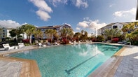 Located at 9500 Grandtree Ave in Venice, Florida, this outdoor amenity space offers a resort-inspired retreat designed for relaxation and leisure. At the heart of the area is a generously sized swimming pool with crystal-clear blue water, framed by a broad paver deck that accommodates rows of modern sling-style lounge chairs ideal for sunbathing. The pool's entry features a gradual shallow zone, making it both accessible and inviting. Surrounding the pool, vibrant tropical landscaping creates a lush, immersive atmosphere, with an abundance of palm trees, red-leafed tropical plants, and dense green foliage that evoke a true Florida resort experience. Paved walkways wind through the greenery, connecting multiple seating and lounging areas, including a shaded pergola structure and intimate outdoor furniture groupings nestled among the palms and colorful plantings. The space is enclosed by a sleek black metal fence, offering privacy and security while maintaining open sightlines to the sky. Multi-story residential buildings frame the backdrop, reinforcing the community setting. Natural light floods the entire area throughout the day, and the combination of lush plantings and open hardscape creates a dynamic, welcoming environment well-suited for outdoor gatherings and quiet relaxation alike.