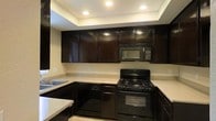 This single-floor space is thoughtfully designed to blend practicality with a clean, modern aesthetic. The kitchen serves as a standout feature, showcasing rich dark wood cabinetry that extends to the ceiling, creating a bold and sophisticated look. Light-toned countertops provide a striking contrast against the dark lower and upper cabinets, offering ample workspace for cooking and meal preparation. The kitchen is fully equipped with matching black appliances, including a gas range with oven, a built-in over-the-range microwave, and a dishwasher, all seamlessly integrated into the cabinetry for a cohesive appearance. A sink with dark cabinetry below completes the functional layout. Recessed lighting is embedded in a subtly elevated ceiling detail above the kitchen, casting a warm, even glow throughout the space and enhancing the overall ambiance. The neutral wall tones complement the dark cabinetry and light countertops, contributing to a balanced and inviting atmosphere. The layout is efficient and well-organized, making excellent use of the available space while maintaining an open and accessible feel throughout.