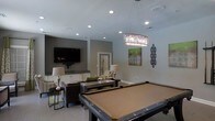 This single-floor entertainment space is thoughtfully designed to serve as a premier social and recreational hub. At the heart of the room sits a full-size billiards table, positioned beneath a striking rectangular crystal chandelier that adds a touch of glamour to the space. The room is bathed in warm, layered lighting, combining recessed ceiling lights with the chandelier's ambient glow to create an inviting atmosphere.

A comfortable lounge area anchors one side of the room, featuring plush seating arranged around a low coffee table — ideal for relaxed conversation or watching the wall-mounted flat-screen television. The TV is set against a tastefully painted accent wall in a deep gray tone, providing a visual contrast to the lighter surrounding walls.

A wet bar or kitchenette area is visible in the background, complete with white cabinetry and dark countertops, offering a convenient space for refreshments. Decorative artwork and wall accents add personality throughout the room. The light-toned tile flooring ties the space together, reflecting light and enhancing the open, airy feel. Overall, the space strikes a balance between sophisticated design and functional entertainment, making it well-suited for social events and leisure activities.
