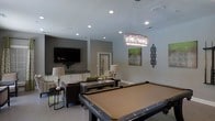 This single-floor entertainment space is thoughtfully designed to serve as a premier social and recreational hub. At the heart of the room sits a full-size billiards table, positioned beneath a striking rectangular crystal chandelier that adds a touch of glamour to the space. The room is bathed in warm, layered lighting, combining recessed ceiling lights with the chandelier's ambient glow to create an inviting atmosphere.

A comfortable lounge area anchors one side of the room, featuring plush seating arranged around a low coffee table — ideal for relaxed conversation or watching the wall-mounted flat-screen television. The TV is set against a tastefully painted accent wall in a deep gray tone, providing a visual contrast to the lighter surrounding walls.

A wet bar or kitchenette area is visible in the background, complete with white cabinetry and dark countertops, offering a convenient space for refreshments. Decorative artwork and wall accents add personality throughout the room. The light-toned tile flooring ties the space together, reflecting light and enhancing the open, airy feel. Overall, the space strikes a balance between sophisticated design and functional entertainment, making it well-suited for social events and leisure activities.