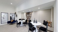 This single-floor business center offers a refined and sophisticated environment designed to support professional work and collaboration. The space is appointed with a clean, contemporary aesthetic, featuring crisp white walls, recessed lighting, and light wood-tone flooring that creates a bright, welcoming atmosphere throughout.

The seating area is thoughtfully arranged with round white pedestal tables paired with ornate black upholstered chairs, striking a balance between modern and classic design sensibilities. A built-in upholstered bench lines one wall, providing additional seating and contributing to the room's polished, lounge-like feel. Decorative black ceramic vases and sculptural accents adorn the tabletops, adding an artistic touch to the functional space.

A dramatic focal point is created by a bold, floor-to-ceiling decorative curtain in gold and navy tones, introducing a rich burst of color and texture against the otherwise neutral palette. Padded wall panels in a soft beige tone further enhance the space's refined character while contributing to a quieter acoustic environment.

The overall ambiance is professional yet inviting, making this business center well-suited for meetings, focused work sessions, or casual networking in an environment that prioritizes both style and comfort.