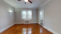 This single-floor space showcases a thoughtfully laid-out floor plan with a warm, classic character throughout. Rich, polished hardwood floors run continuously across the living and bedroom areas, reflecting natural light and adding a timeless appeal. Crown molding accents the ceilings in several rooms, lending an air of traditional elegance, while ceiling fans provide both function and style.

The living areas are generously proportioned, with neutral gray walls creating a versatile backdrop. Multiple windows, including a bay window configuration in one room, allow ample natural light to fill the space and offer views of the surrounding neighborhood. Classic radiator units are present beneath several windows, underscoring the building's period character.

The kitchen features white cabinetry with ample storage, dark countertops, and tile flooring. It is equipped with white appliances including a refrigerator, range, microwave, and dishwasher, providing a fully functional cooking space.

The bathroom is straightforward and clean, with tile flooring, a single vanity with storage, a mirror cabinet, and a window for natural ventilation. Throughout the unit, wide baseboards and white trim details add a cohesive, classic finish that ties each room together into a comfortable and characterful living space.