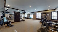This fitness center is a thoughtfully designed space that offers a comprehensive workout environment within a single open floor plan. The room is finished with warm beige walls, dark wood crown molding and baseboards, and wall-to-wall carpeting, lending a polished yet functional aesthetic throughout.

The cardio zone is well-stocked with multiple treadmills, elliptical trainers, a stationary bike, and a stair climber, each positioned to take advantage of the natural light streaming through several large, multi-pane windows along the exterior wall. Mounted televisions provide entertainment during workouts. A dedicated strength training area features a multi-station cable machine, an adjustable weight bench, and a full dumbbell rack with a wide range of colorful free weights, accommodating a variety of fitness routines.

A floor-to-ceiling mirrored wall section visually expands the space and supports stretching and form-checking exercises. Water coolers are conveniently positioned nearby. Recessed lighting throughout the ceiling ensures even, bright illumination at all hours. Framed artwork and a wall clock add subtle decorative touches. The overall atmosphere is clean, organized, and conducive to focused exercise.