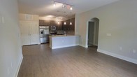 This single-floor residence showcases an open-concept layout that seamlessly connects the living area and kitchen, creating a spacious and functional environment. The hardwood-style flooring extends throughout, providing a warm, cohesive foundation that ties the space together.

The kitchen is a standout feature, equipped with stainless steel appliances, including a French door refrigerator and microwave. Dark espresso-toned cabinetry lines the walls, offering generous storage, while a central white island with a light-toned countertop provides additional prep space and serves as a natural dividing point between the kitchen and living area. Pendant lighting hangs above the island, complementing the track lighting overhead to ensure the space is well-lit.

The open living area is generously proportioned, offering flexibility in furniture arrangement. An elegant arched doorway adds an architectural detail that leads to additional areas of the residence. Neutral wall tones throughout contribute to a clean, modern aesthetic. A white-paneled door near the kitchen provides access to an entry or utility area, completing this well-appointed, move-in ready space.
