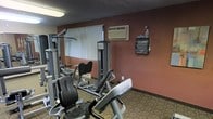 This single-floor fitness room offers a functional workout space equipped with a selection of strength-training and cardio machines. The room features warm terracotta-toned walls paired with patterned carpeting, creating a utilitarian yet welcoming atmosphere. A wall-mounted air conditioning unit provides climate control, ensuring comfort during workouts. Natural light filters in through a vertical-blind-covered window, supplemented by overhead fluorescent lighting that keeps the space well-lit. The strength equipment includes a multi-station cable machine and a seated weight machine, while cardio options such as a stationary bike are also present. A wall-mounted paper towel dispenser and a bottle of sanitizing spray are conveniently positioned, promoting hygiene and cleanliness throughout the space. The textured ceiling adds a classic finishing touch to the room's overall design. Though modest in scale, the fitness room is thoughtfully arranged to maximize the available floor space, making it a practical option for those looking to maintain their exercise routine.