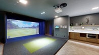 Located at 460 North Broad Street in Philadelphia, this dedicated indoor golf simulator suite delivers a sophisticated, amenity-rich experience within a thoughtfully designed single-room layout. At its centerpiece is a Full Swing golf simulator, complete with a ceiling-mounted projector, professional-grade impact screen, and an artificial turf hitting mat set against a dark carpet surround — creating an immersive, stadium-like atmosphere with dramatic blue accent lighting.

The space transitions seamlessly from the simulator bay to a refined lounge and service area. A built-in wooden cabinetry unit with a white countertop lines one wall, serving as a bar and equipment station, accented by open shelving with greenery and decorative golf accessories. A wall-mounted flat-screen display and informational panels add functional detail to the area.

The lounge corner features a plush green velvet curved sofa and leather chairs, set against a painted grey brick accent wall — a stylish backdrop complemented by mounted sports balls as wall art. Underfoot, a warm herringbone hardwood floor anchors the lounge zone, contrasting beautifully with the grey carpet of the simulator area.

Wall-mounted golf club racks provide organized club storage along the grey brick wall, adding both practicality and visual interest. Recessed lighting and a central circular HVAC vent complete the polished, well-appointed ceiling. This suite offers a complete, club-quality golf and entertainment experience.