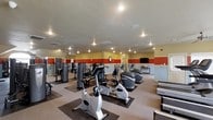 Located at 21 Player Ln in Starkville, Mississippi, this fitness center offers a comprehensive workout facility designed to accommodate a variety of exercise routines. The single-floor space is outfitted with an extensive selection of equipment, including stationary bikes, elliptical machines, and a range of strength training stations, providing users with options for both cardiovascular and resistance-based workouts. The interior features a neutral carpet flooring with rubber equipment mats, recessed lighting throughout the ceiling, and a bold two-tone wall design with warm yellow and red accent bands that create an energizing atmosphere. Wall-mounted mirrors along one side of the room enhance the sense of space and allow exercisers to monitor their form. A wall-mounted flat-screen television is positioned for convenient viewing during workouts, and a wall clock keeps members on schedule. The space is well-lit with recessed ceiling fixtures and benefits from a clean, organized layout that allows for easy movement between stations. Subtle ventilation and fire safety systems are integrated into the ceiling, reflecting attention to comfort and safety throughout the facility.