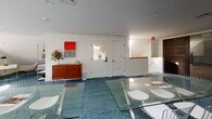 The Community Room at 703 Whitney Ave in New Haven, Connecticut is a thoughtfully designed, single-floor gathering and meeting space that blends modern aesthetics with functional versatility. The room is characterized by its clean white walls and angled ceiling lines that lend an airy, loft-like quality to the interior. Abundant natural light streams in through multiple skylights, casting bright pools of light across the rich teal-blue carpeted floors and creating a welcoming atmosphere throughout the day. At the heart of the space sits a long glass-top conference table with a sleek chrome base, surrounded by modern wire-frame chairs — an arrangement well-suited for meetings, workshops, or collaborative gatherings. A dedicated alcove area features a minimalist white desk with a stylish bronze desk lamp, offering a more private workspace for individual use. Decorative touches include a mid-century walnut dresser, a bold red abstract artwork, and a slender open bookshelf that add warmth and personality to the otherwise crisp, contemporary palette. Access to an adjacent area is provided through a set of panel doors, with a staircase visible beyond — connecting the room to the broader building. Windows along one wall allow additional natural light to filter in, reinforcing the space's open, productive ambiance.
