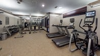 This fitness center offers a thoughtfully organized layout designed to accommodate a full range of workout routines. The space is finished with neutral-toned walls, dark wood crown molding and baseboards, and low-pile carpet flooring that provides both comfort and durability underfoot.

The cardio zone features multiple treadmills and elliptical machines, each equipped with individual digital display consoles. Full-length mirrors line the walls, visually expanding the space and allowing users to monitor their form throughout their workout.

The strength-training area is outfitted with a selection of cable and resistance machines, including leg extension and curl stations, a multi-function cable tower, and additional functional training equipment. A stability ball rounds out the accessories available for core and flexibility work.

Recessed and overhead lighting illuminates the space evenly, creating a bright and energizing environment. The ceiling height contributes to an open, airy feel throughout. The overall design is clean and functional, with a classic aesthetic reinforced by the consistent use of dark wood trim accents. The fitness center is well-maintained and provides users with all the essentials needed for both cardiovascular conditioning and strength development in a focused, professional setting.