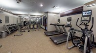 This fitness center offers a thoughtfully organized layout designed to accommodate a full range of workout routines. The space is finished with neutral-toned walls, dark wood crown molding and baseboards, and low-pile carpet flooring that provides both comfort and durability underfoot.

The cardio zone features multiple treadmills and elliptical machines, each equipped with individual digital display consoles. Full-length mirrors line the walls, visually expanding the space and allowing users to monitor their form throughout their workout.

The strength-training area is outfitted with a selection of cable and resistance machines, including leg extension and curl stations, a multi-function cable tower, and additional functional training equipment. A stability ball rounds out the accessories available for core and flexibility work.

Recessed and overhead lighting illuminates the space evenly, creating a bright and energizing environment. The ceiling height contributes to an open, airy feel throughout. The overall design is clean and functional, with a classic aesthetic reinforced by the consistent use of dark wood trim accents. The fitness center is well-maintained and provides users with all the essentials needed for both cardiovascular conditioning and strength development in a focused, professional setting.