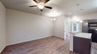 This single-story space offers a thoughtfully designed open-concept layout that seamlessly connects the main living and dining areas. The living room features durable wood-look hardwood flooring, neutral light gray walls, and a ceiling fan with integrated lighting, creating a comfortable and versatile space. A chandelier marks the dining area, which flows naturally from the living room toward the kitchen.

The kitchen is a functional highlight, featuring dark gray cabinetry with ample storage, light granite-style countertops, a peninsula with pendant lighting overhead, and black appliances including a range, microwave, dishwasher, and refrigerator. The wood-look flooring continues throughout this area, maintaining a cohesive aesthetic.

The bedrooms are finished with plush carpeting and ceiling fans, providing a quieter, comfortable atmosphere. Natural light filters in through window blinds, adding warmth to each room. One bedroom connects to a hallway leading to additional spaces within the unit.

The bathroom is clean and straightforward, featuring a full tub-shower combo with built-in shelving, wood-look flooring, dark cabinetry, and granite-style countertops that coordinate with the kitchen finishes. Overall, the space maintains a consistent, modern aesthetic throughout.