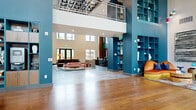 This thoughtfully designed communal space makes a striking first impression through its grand lobby, where soaring double-height ceilings and an expansive wall of clerestory windows flood the interior with natural light. Warm hardwood flooring anchors the open floor plan, while bold teal and green accent walls punctuate the architecture with vibrant color. A backlit wood reception desk commands attention at the center of the lobby, framed by a dramatic floor-to-ceiling teal screen installation and flanked by built-in display shelving curated with decorative objects.

The lounge area offers comfortable seating arrangements set atop layered area rugs, with an upper-level mezzanine visible overhead, adding to the vertical drama of the space. A dedicated business and co-working nook features a striking blue resin table paired with coordinating chairs, built-in wood shelving, and a playful collage-style display wall.

Private meeting rooms are finished with deep green walls, tall windows offering garden views, dark marble-effect tile flooring, and mid-century modern wood chairs surrounding a green conference table. Corridors are detailed with veined marble-look tile and original artwork. A dedicated mail room provides rows of individual mailboxes and package lockers, finished with polished granite-clad partitions.