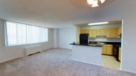 This single-floor space features an open-concept design that seamlessly connects the living area and kitchen, creating a cohesive and functional layout. The spacious living area is finished with wall-to-wall carpeting in a neutral tone, contributing to a clean and understated aesthetic. A large window dressed with vertical blinds allows generous natural light to fill the room, enhancing its open feel. A baseboard heating unit runs along the exterior wall beneath the window, providing practical climate control.

The kitchen is partially separated from the living area by a peninsula counter finished with a dark laminate surface, offering additional workspace and a casual dining or prep area. Light wood cabinetry lines the kitchen walls, providing ample storage, while under-cabinet fluorescent lighting illuminates the workspace effectively. Black appliances, including a full-sized refrigerator, are integrated into the layout. The kitchen flooring transitions to tile, clearly delineating the cooking space from the carpeted living area. Overhead, a ceiling fan with a light fixture provides both ventilation and ambient lighting throughout the open space.