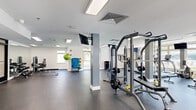 This fitness center offers a spacious, open-plan layout across a single floor, thoughtfully designed to accommodate a full range of workout activities. The space is finished with durable speckled rubber flooring throughout, providing both safety and comfort underfoot. Structural columns painted in a clean gray tone help define zones within the open layout without interrupting the overall flow of the space.

The cardio section lines the perimeter near large windows, housing multiple treadmills and elliptical machines, all positioned to take advantage of the ample natural light and outdoor views. Wall-mounted flat-screen televisions are strategically placed throughout, keeping users engaged during workouts. A dedicated strength training area features a multi-station cable machine and free weight equipment, including benches and weight racks by True Fitness.

A separate functional training zone offers open floor space along with stability balls, resistance equipment, and modular storage for accessories. This area benefits from direct outdoor access and additional window views. A wall of mirrors enhances the sense of space and supports form-checking during exercise.

The entry area includes a stainless steel hydration station, fire safety equipment, and double glass exit doors. Skylight panels in the ceiling supplement the natural light from the surrounding windows, creating a bright and energizing atmosphere throughout the facility.