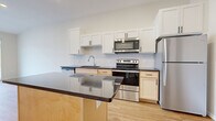 This single-floor unit showcases a clean, contemporary design highlighted by light hardwood flooring that runs throughout the open living areas. The kitchen serves as a central feature, combining functionality and style with a full suite of stainless steel appliances, including a refrigerator, electric range, built-in microwave, and dishwasher. White shaker-style upper cabinets contrast neatly with warm natural wood lower cabinets, while a white subway tile backsplash adds a polished finish. Dark countertops extend across the kitchen and onto a freestanding island, which is illuminated by pendant lighting above—creating a natural gathering point within the open-plan layout. The living area is bright and airy, with tall ceilings and large sliding glass doors that lead to a private balcony equipped with metal railings, offering views of the surrounding community. The bathroom features a light wood vanity with ample storage, a white countertop, and a wide mirror. Neutral tones and recessed lighting throughout contribute to a cohesive, modern ambiance that is both inviting and practical.