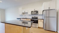 This single-floor unit showcases a clean, contemporary design highlighted by light hardwood flooring that runs throughout the open living areas. The kitchen serves as a central feature, combining functionality and style with a full suite of stainless steel appliances, including a refrigerator, electric range, built-in microwave, and dishwasher. White shaker-style upper cabinets contrast neatly with warm natural wood lower cabinets, while a white subway tile backsplash adds a polished finish. Dark countertops extend across the kitchen and onto a freestanding island, which is illuminated by pendant lighting above—creating a natural gathering point within the open-plan layout. The living area is bright and airy, with tall ceilings and large sliding glass doors that lead to a private balcony equipped with metal railings, offering views of the surrounding community. The bathroom features a light wood vanity with ample storage, a white countertop, and a wide mirror. Neutral tones and recessed lighting throughout contribute to a cohesive, modern ambiance that is both inviting and practical.