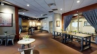 This single-floor commercial space offers a dynamic and versatile environment well-suited for a variety of events and gatherings. The open-plan layout is anchored by a mix of seating options, including communal long tables with bench seating, small bistro-style round tables, and bar-height chairs, providing flexibility for both intimate and larger group settings.

The interior design strikes an eclectic industrial tone, with exposed brick walls, textured concrete columns, and rich burgundy accent walls that add warmth and character. A drop tile ceiling with recessed and track lighting provides even, adaptable illumination throughout the space.

Toward the back, a dedicated bar and kitchenette area is fitted with cabinetry, counter space, and additional bar seating, making it well-equipped for catered events or beverage service. A dramatic draped curtain partition near the entrance offers the option to section off areas, adding a layer of privacy or staging capability.

Framed artwork adorns the walls, contributing to an inviting and curated ambiance. The carpeted flooring in warm, earthy tones adds comfort underfoot, while the open sightlines throughout the space make it easy to navigate. Overall, this space combines a welcoming atmosphere with practical amenities, making it a capable venue for corporate functions, private parties, or social events.