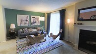 This single-floor residence offers a thoughtfully designed living space that balances comfort with contemporary style. The living room serves as the centerpiece of the home, featuring a striking green accent wall adorned with artistic wall décor, including bicycle-themed prints that add a creative, urban touch. A large window dressed with flowing dark curtains and sheer panels floods the room with natural light, creating an open and airy ambiance. The space is furnished with a plush sectional sofa complemented by stylish butterfly chairs, all anchored by a vibrant geometric-patterned area rug that adds a lively pop of color to the carpeted floor. Multiple floor lamps provide warm ambient lighting throughout the room. The textured ceiling adds a classic residential character to the space. The overall design aesthetic blends modern and eclectic elements, resulting in a cozy yet visually dynamic environment well-suited for both relaxing and entertaining.